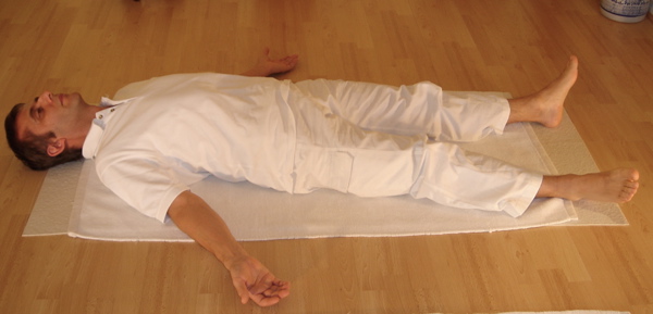 man in white doing corpse pose on mat on wooden floor
