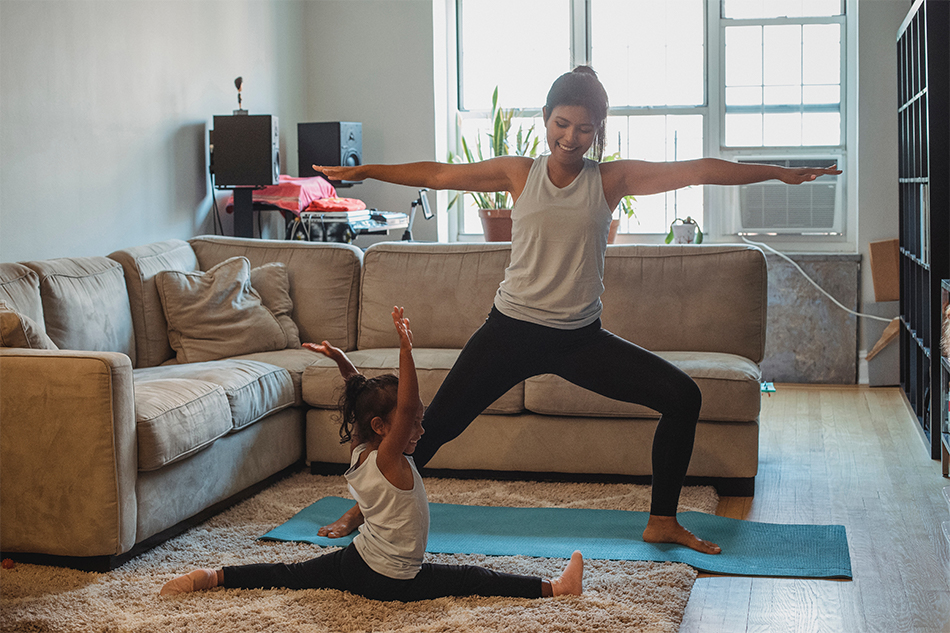young woman performing warrior pose in living room with daughter nearby doing splits